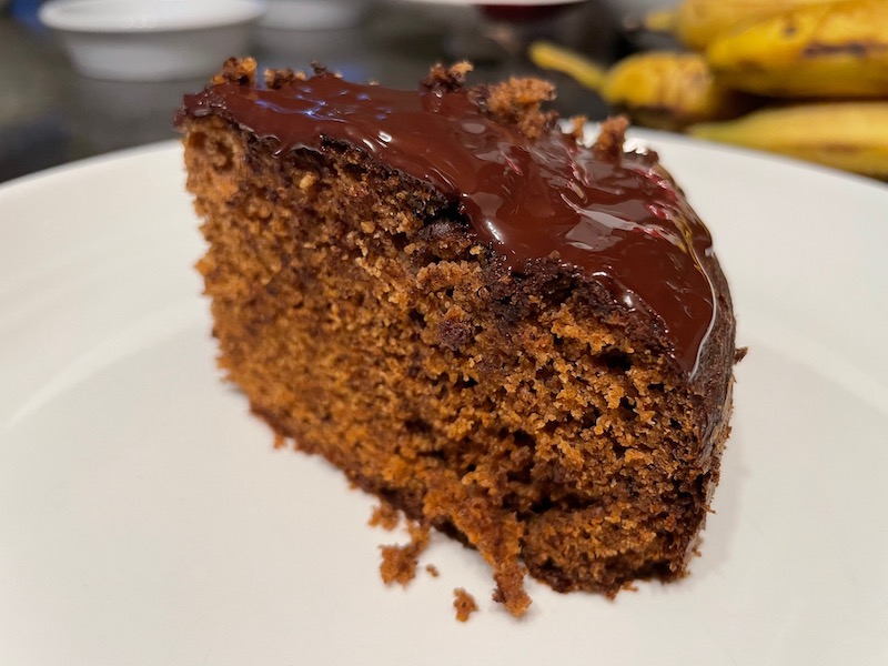 A photograph of a slice of banana bread sitting on a white plate, with a few bananas visible in the blurry background. The slice has a top layer of chocolate, making it look like a cake.